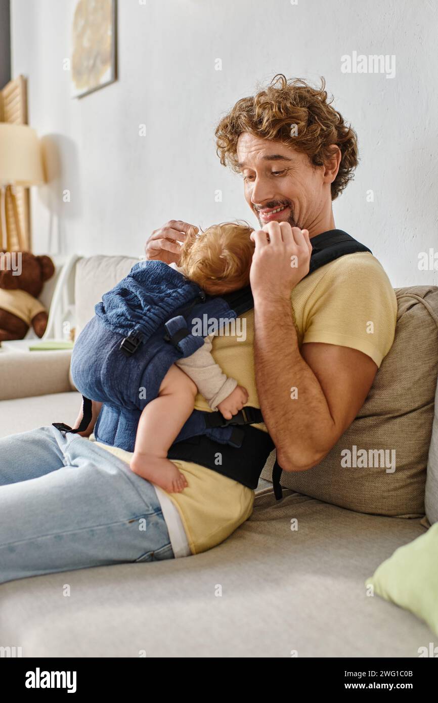 Fröhlicher, lockiger Vater, der auf der Couch sitzt, mit dem Kleinkind in Babytrage, Vaterschaft und Liebe Stockfoto