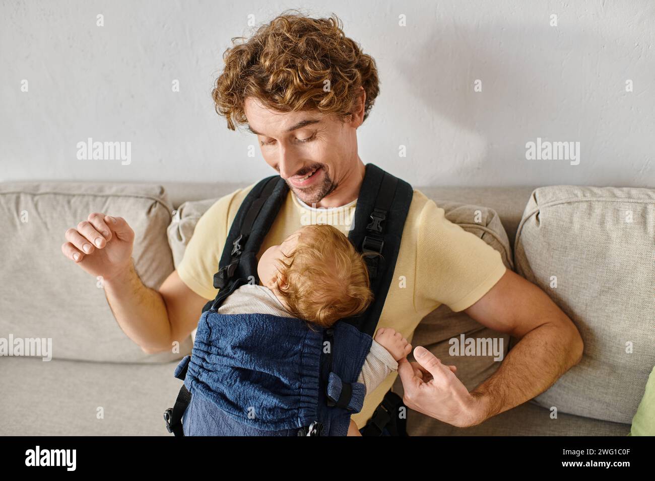 Fröhlicher Mann mit lockigen Haaren, der seinen kleinen Sohn in Babytrage, Vaterschaft und Liebe ansieht Stockfoto