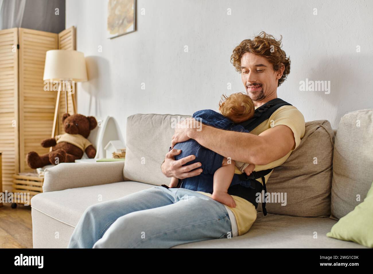 Glücklicher Vater mit lockigen Haaren, der auf der Couch sitzt, mit einem kleinen Sohn in Babytrage, Vaterschaft und Liebe Stockfoto