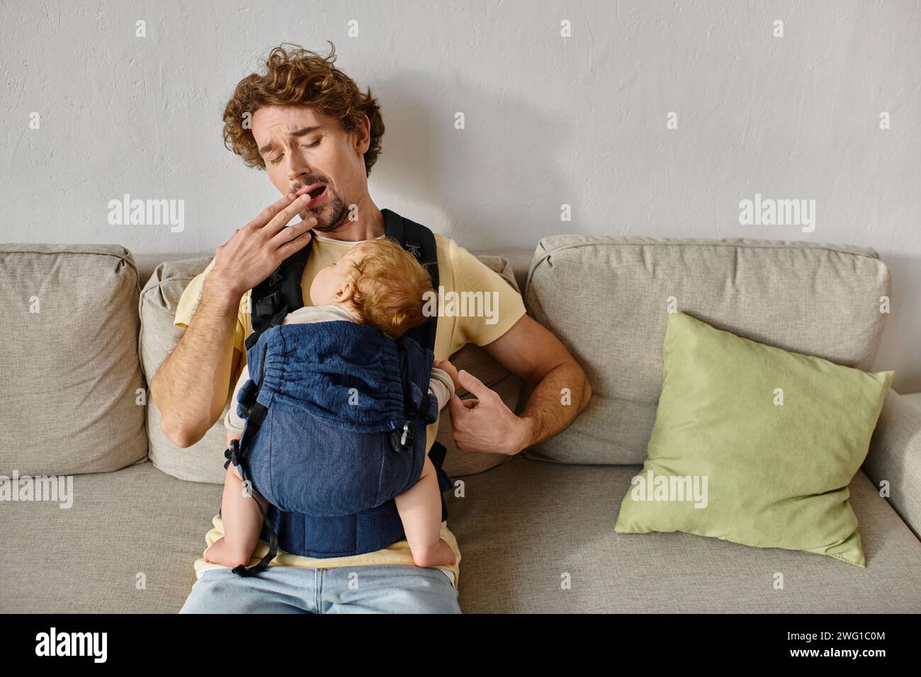 Müder Mann mit lockigen Haaren gähnt mit Kleinkind Sohn in Babytrage im Wohnzimmer, Vaterschaft und Liebe Stockfoto