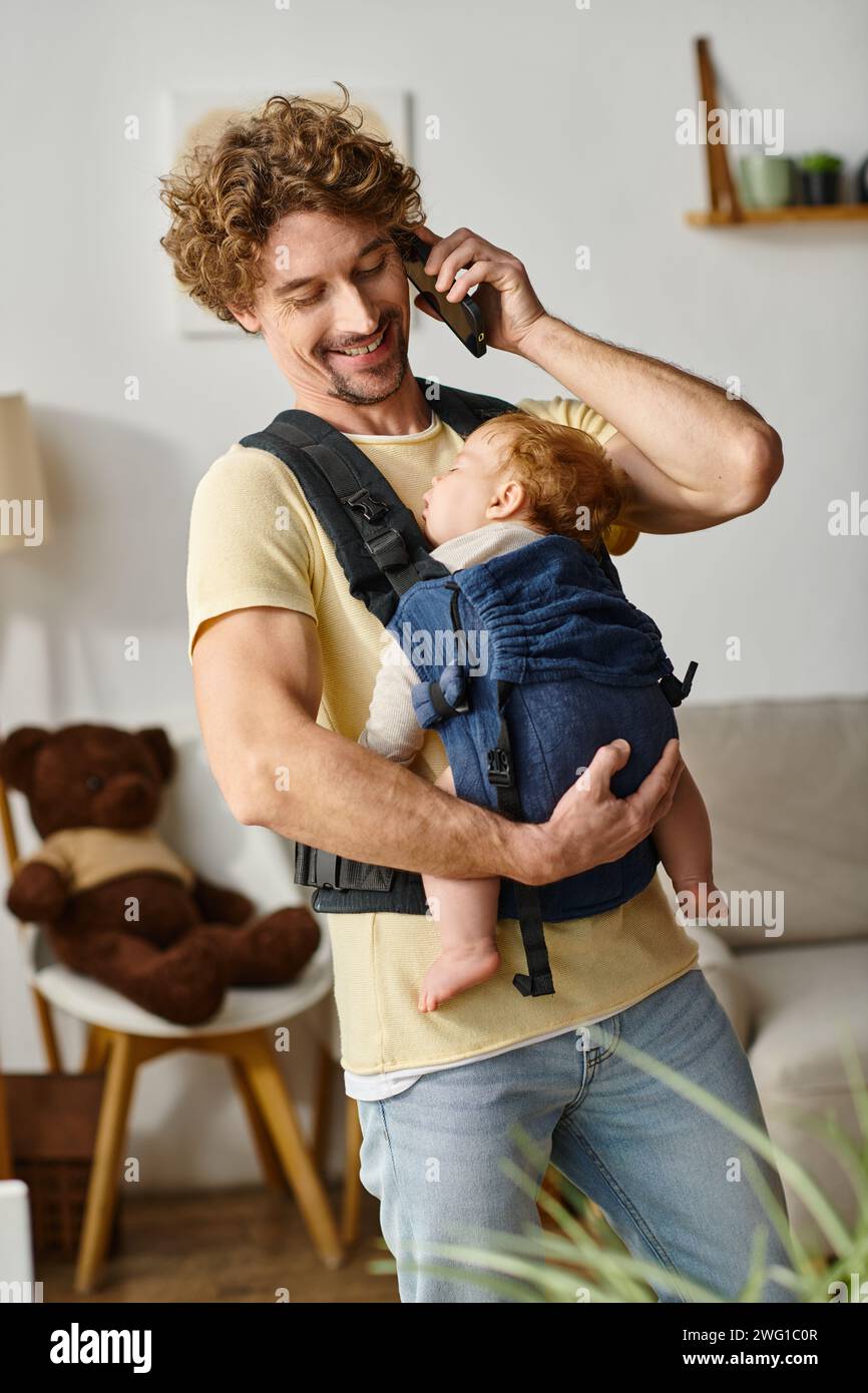 Fröhlicher Vater, der auf dem Smartphone spricht, während das Baby in der Tragetasche schläft, Balance zwischen Leben und Arbeit Stockfoto