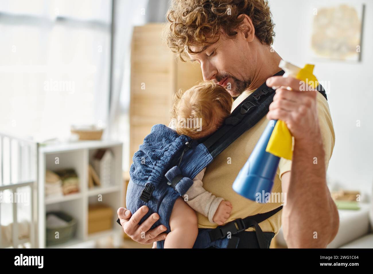 Glücklicher Vater küsst den verschlafenen Sohn in der Tragetasche und hält Sprühflasche und Lappen, Hausarbeit Stockfoto