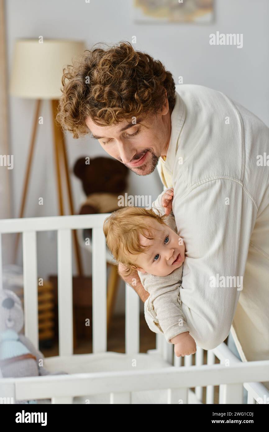 Liebevoller lockiger Vater, der seinen kleinen Sohn mit blauen Augen aus der Kinderkrippe hebt, Vaterschaft Stockfoto