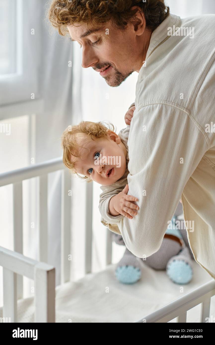 Der liebevolle und glückliche Vater hebt seinen kleinen Sohn mit blauen Augen aus der Kinderkrippe, Vaterschaft Stockfoto