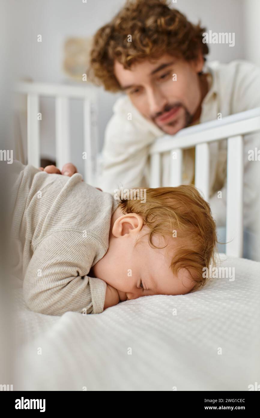 Niedlicher kleiner Junge, der in der Krippe schläft, während der Vater ihn beobachtet, alleinerziehender Vater auf verschwommenem Hintergrund Stockfoto