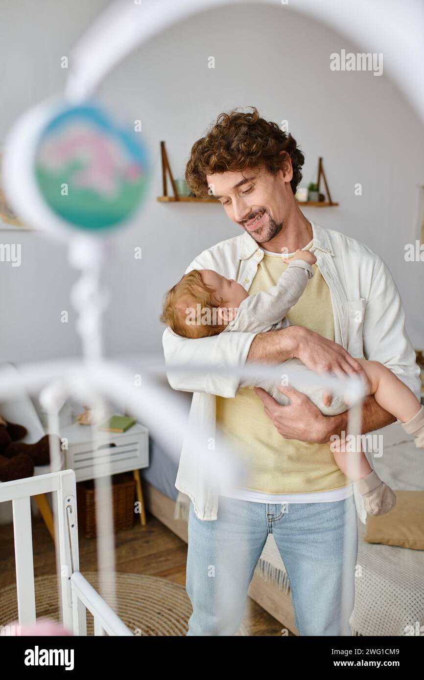 Glücklicher alleinerziehender Vater, der seinen kleinen Sohn in Armen in der Nähe einer Krippe hält, in Kinderzimmer, Vaterschaft und Liebe Stockfoto
