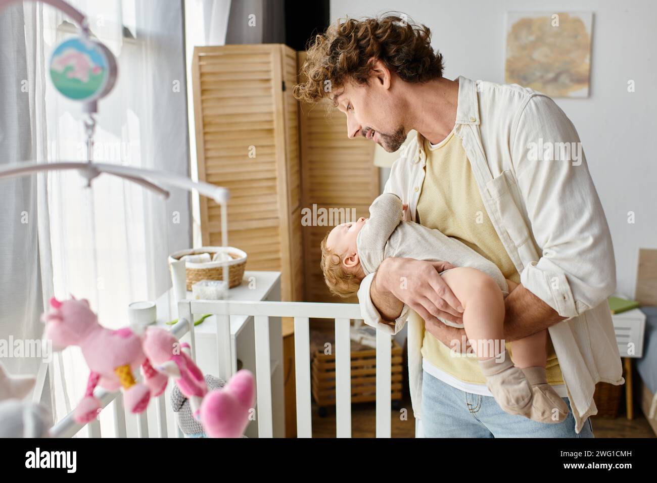 Fröhlicher alleinerziehender Vater, der seinen kleinen Sohn in Armen in der Nähe einer Babykrippe hält, in Kinderzimmer, Vaterschaft und Liebe Stockfoto