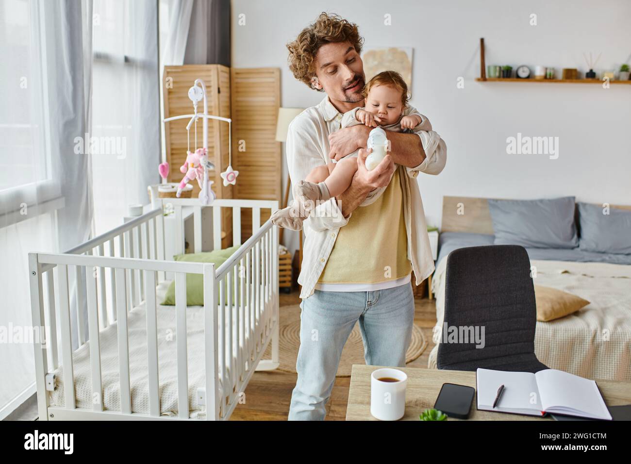 Liebevoller alleinerziehender Vater, der den Säugling aus der Babyflasche ernährt, während er neben dem Kinderbett und dem Schreibtisch steht Stockfoto