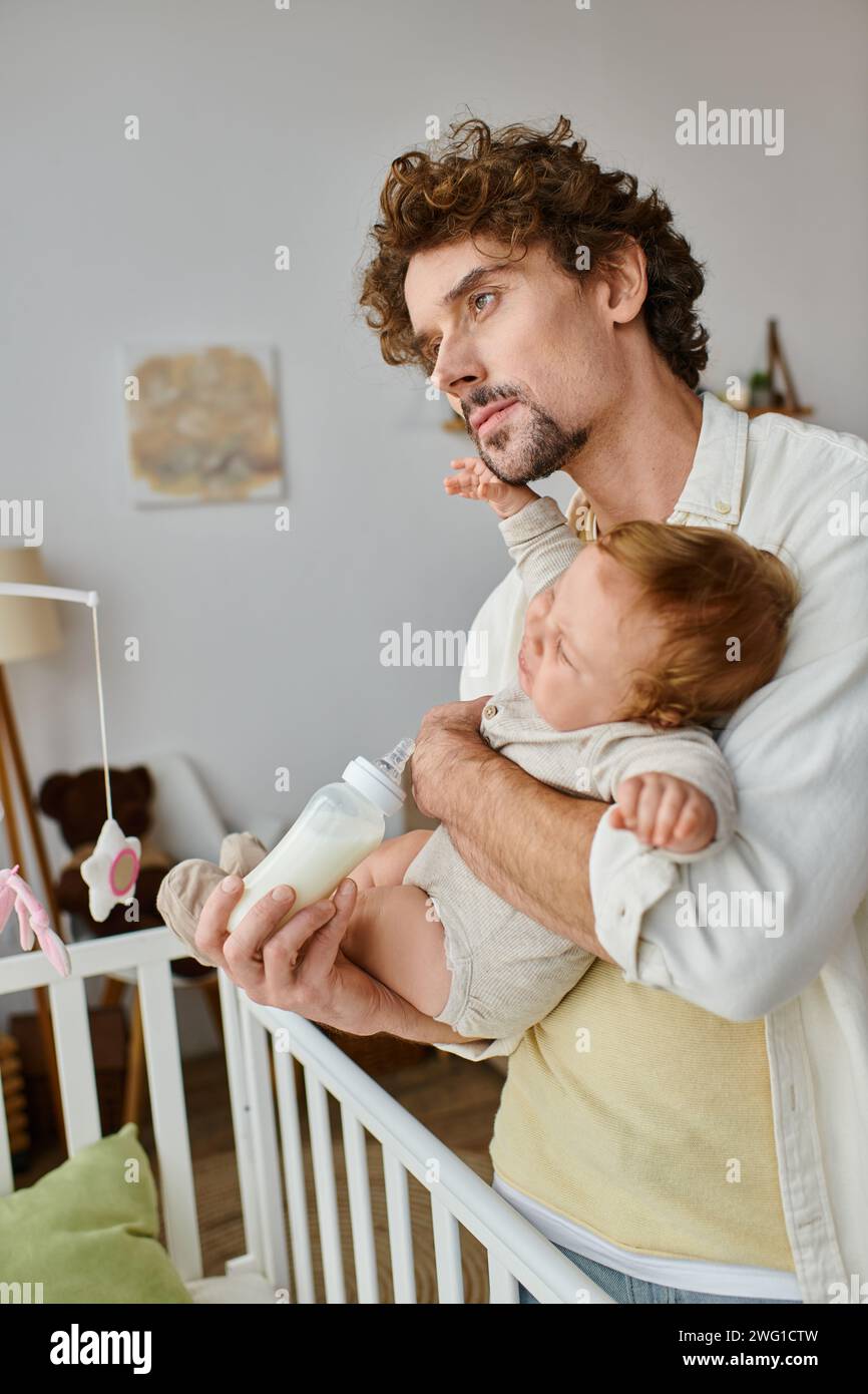 Vater mit lockigen Haaren, der seinen kleinen Sohn aus einer Babyflasche in der Nähe von Kinderbett, Vaterschaft und Pflege ernährt Stockfoto