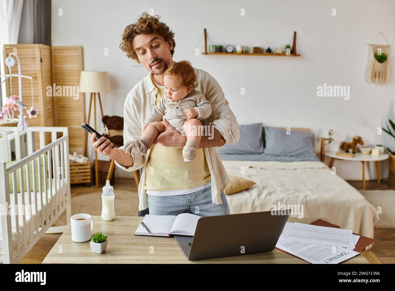 Viel beschäftigter Alleinvater hält den Kleinkind fest und benutzt das Smartphone in der Nähe des Laptops und die Babyflasche auf dem Schreibtisch Stockfoto