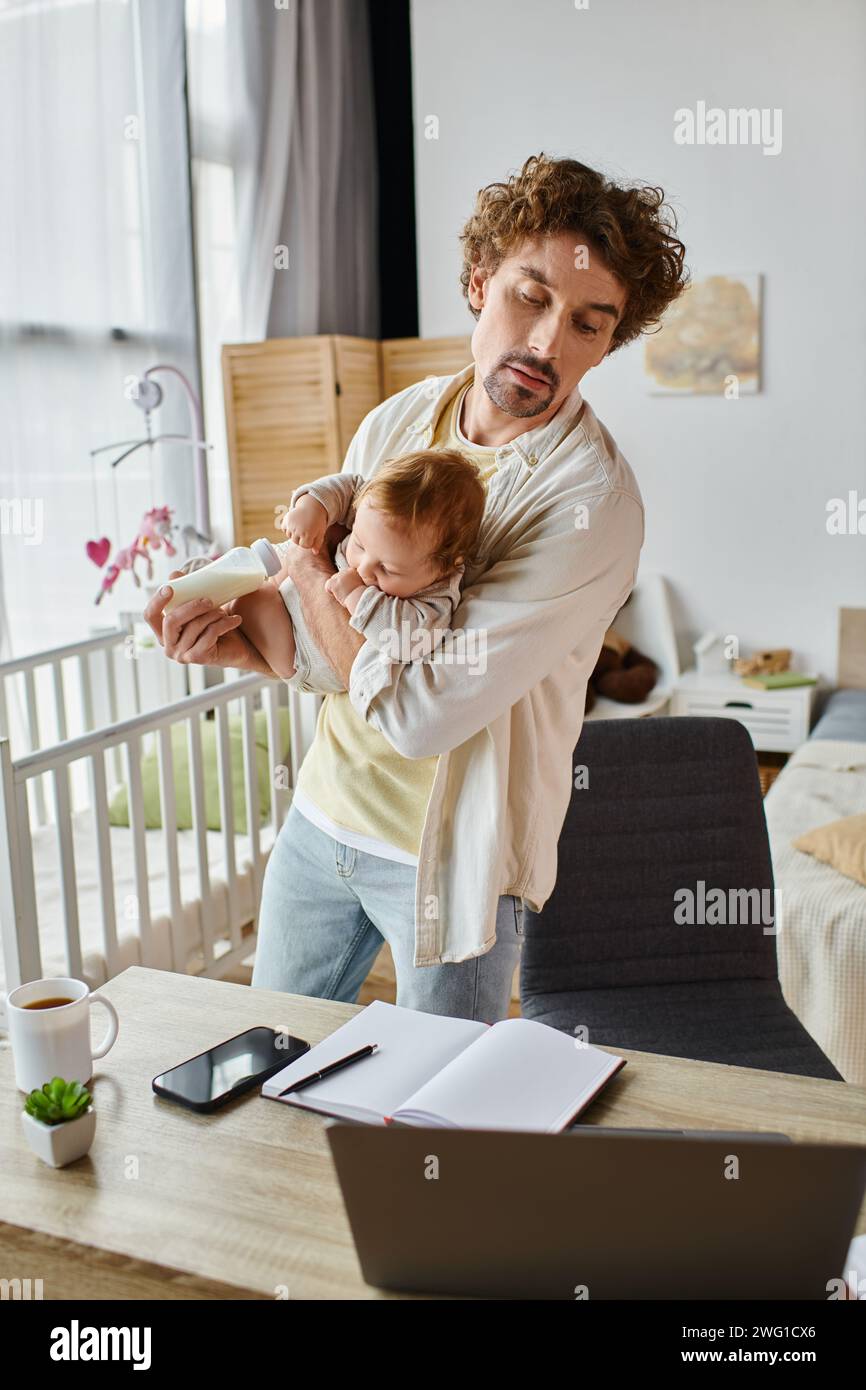 Alleinerziehender Vater hält Sohn und Babyflasche, während er auf den Laptop auf dem Schreibtisch blickt, Work Life Balance Stockfoto