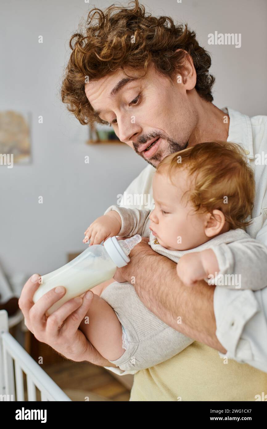 Vater mit lockigen Haaren füttert seinen kleinen Sohn aufmerksam mit Milch in der Babyflasche, Vaterschaft und Pflege Stockfoto