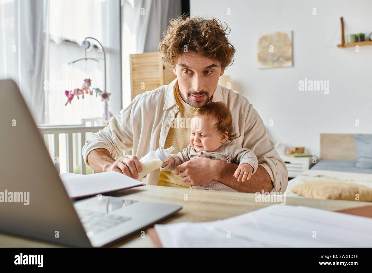 Ein beschäftigter Vater hält die Babyflasche mit Milch und füttert den Sohn des Säuglings, während er von zu Hause aus am Projekt arbeitet Stockfoto