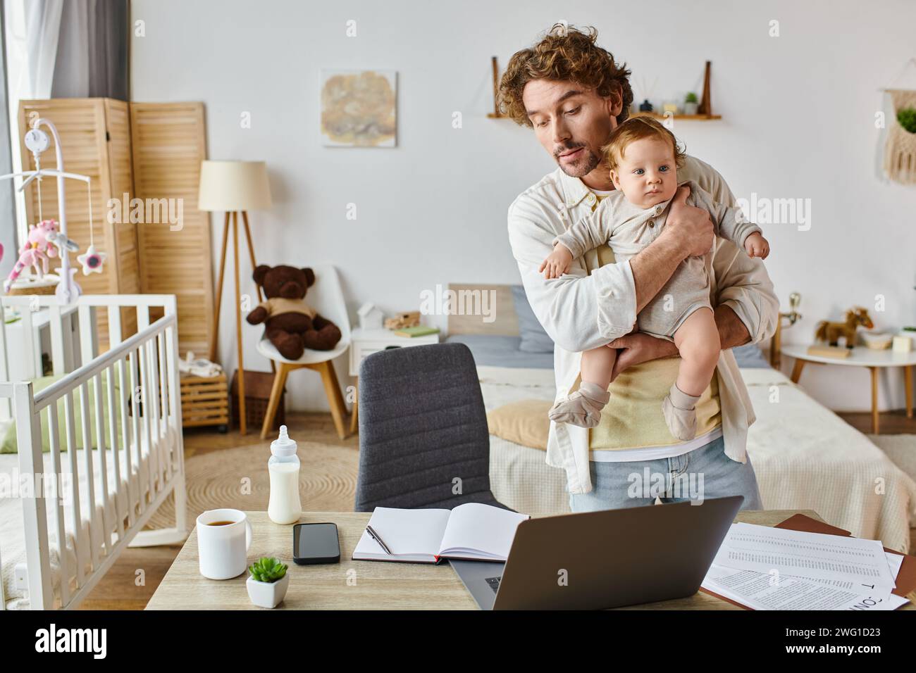Viel beschäftigter alleinstehender Vater hält den Säugling fest und sieht sich Papiere in der Nähe von Gadgets und Babyflasche auf dem Schreibtisch an Stockfoto
