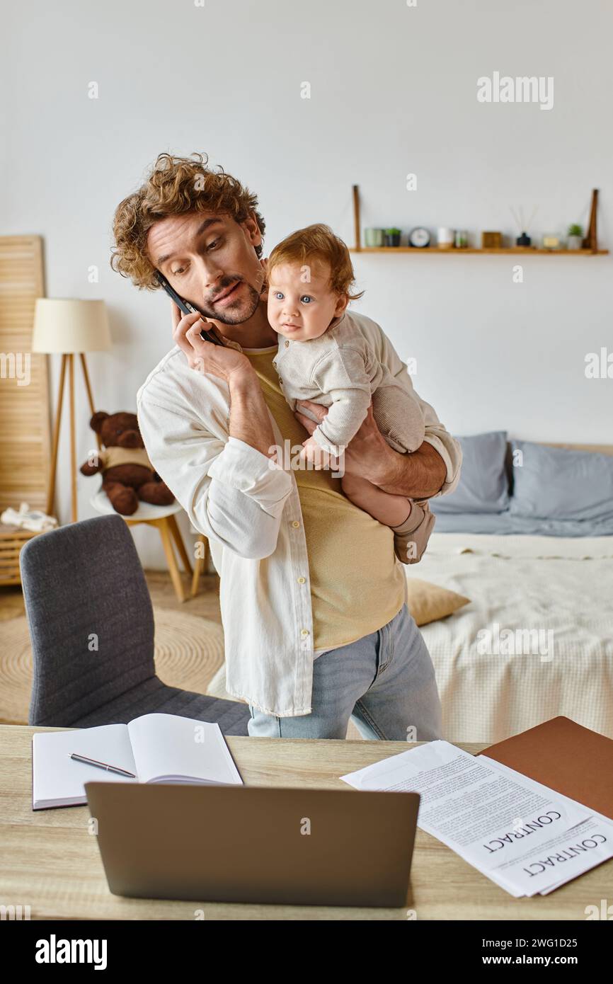 Alleinerziehender Vater hält den Kleinkind fest, während er mit dem Smartphone in der Nähe des Laptops auf dem Schreibtisch spricht, Fernarbeit Stockfoto