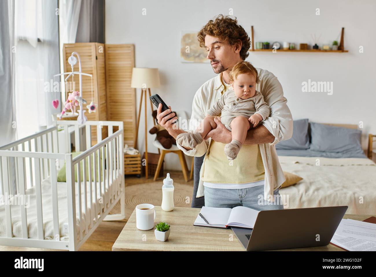 Viel beschäftigter alleinstehender Vater hält den Kleinkind fest und benutzt das Smartphone in der Nähe des Laptops und die Babyflasche auf dem Schreibtisch Stockfoto