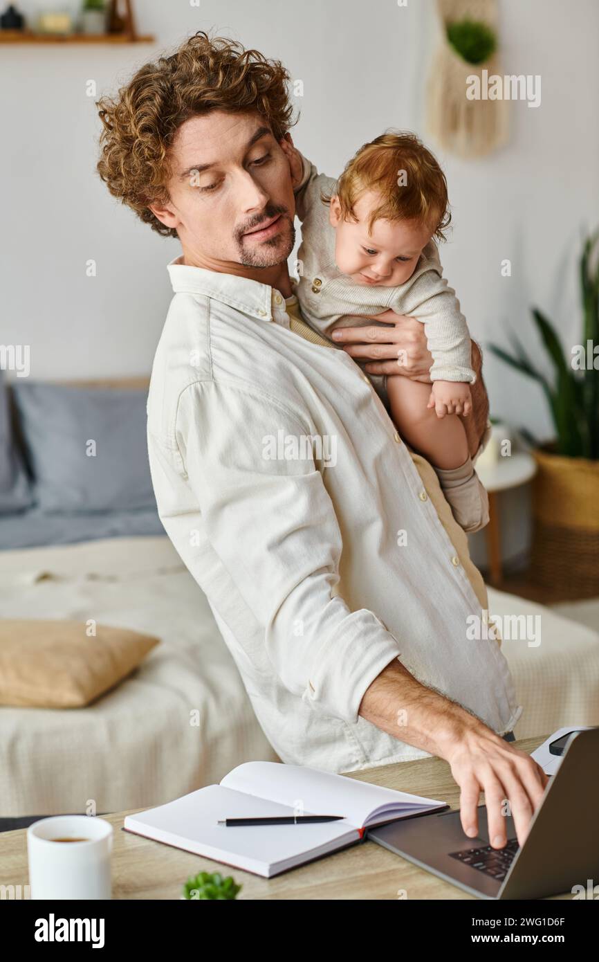 Glücklicher und lockiger Vater, der seinen kleinen Sohn in den Armen hält und mit dem Laptop auf dem Schreibtisch arbeitet und das Leben ausgleicht Stockfoto