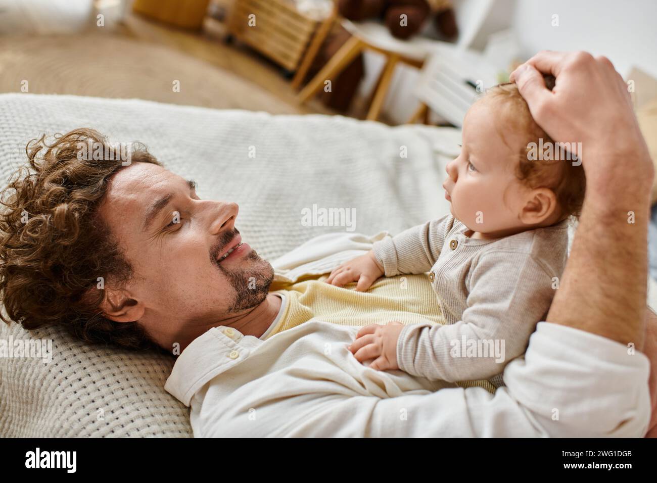 Glücklicher Mann mit lockigen Haaren und Bart, der Haare streichelt, mit seinem kleinen Sohn im Schlafzimmer, Liebe und Pflege Stockfoto