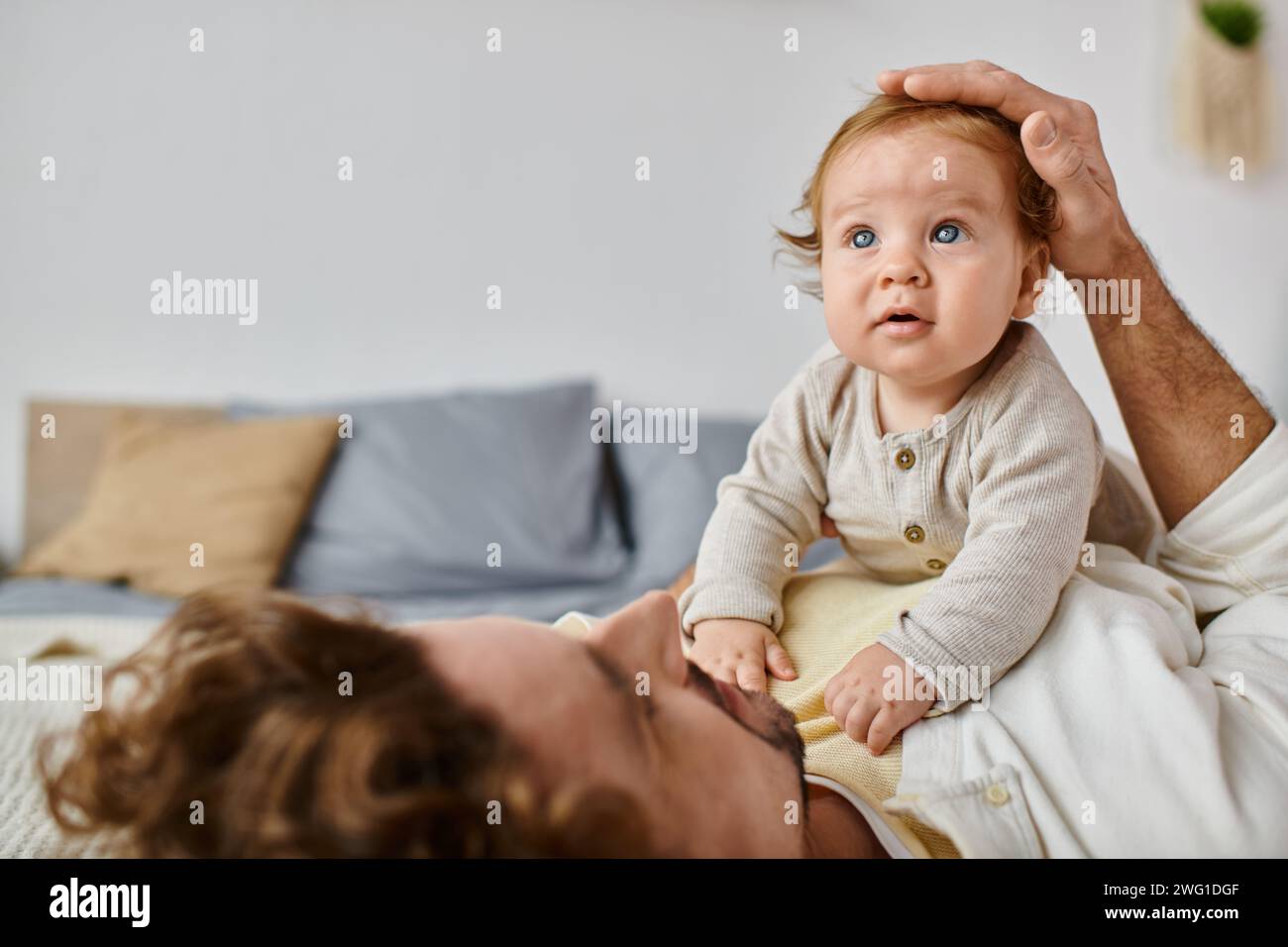 Mann mit lockigen Haaren und Bart streichelt Haare mit seinem kleinen Sohn mit blauen Augen im Schlafzimmer Stockfoto