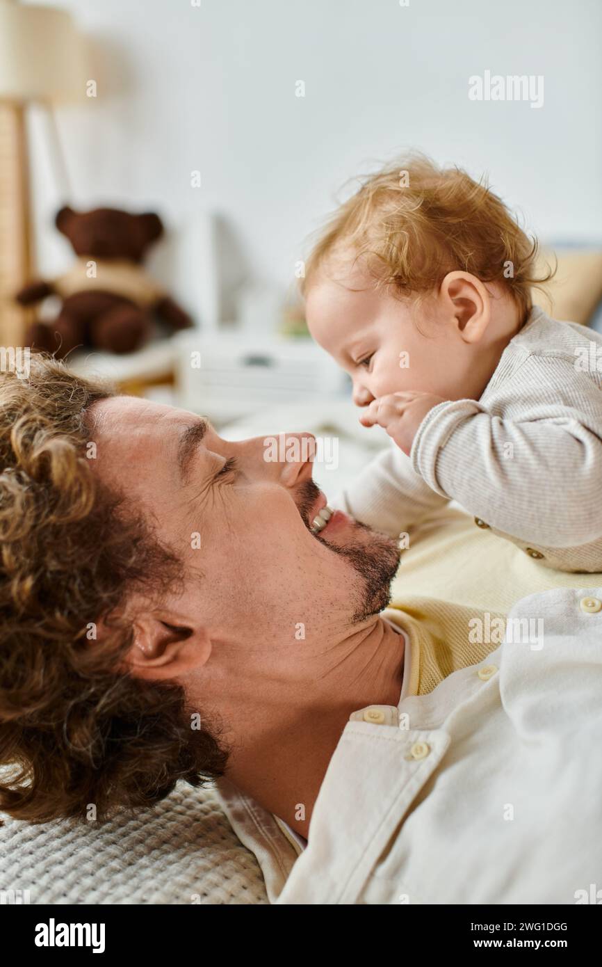 Seitenansicht eines glücklichen Mannes mit lockigen Haaren und Bart, der seinen Sohn im Schlafzimmer ansieht, Liebe und Pflege Stockfoto