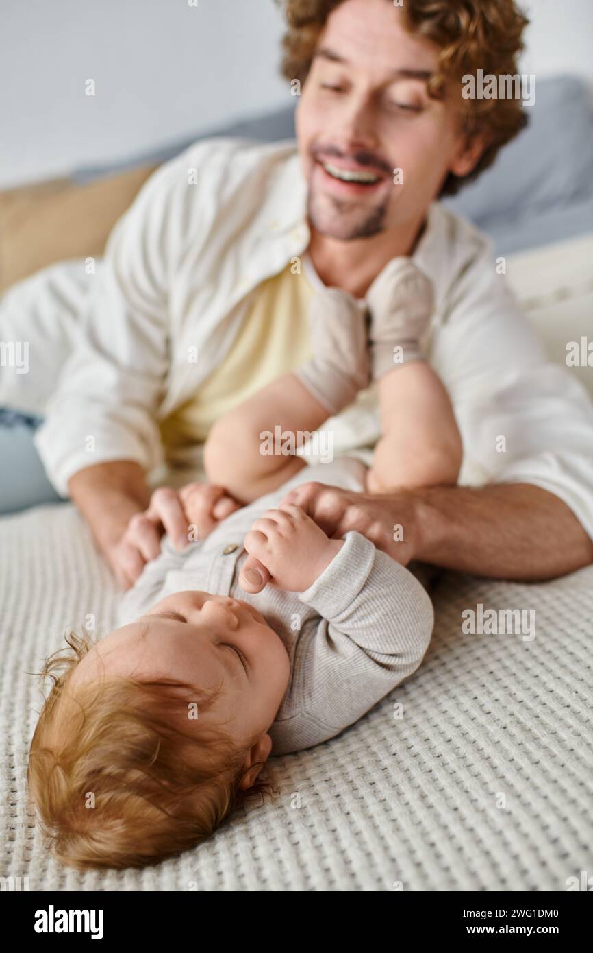 Glücklicher Vater mit lockigen Haaren, der mit seinem kleinen Jungen spielt, während er zusammen auf dem Bett liegt, unschuldig Stockfoto
