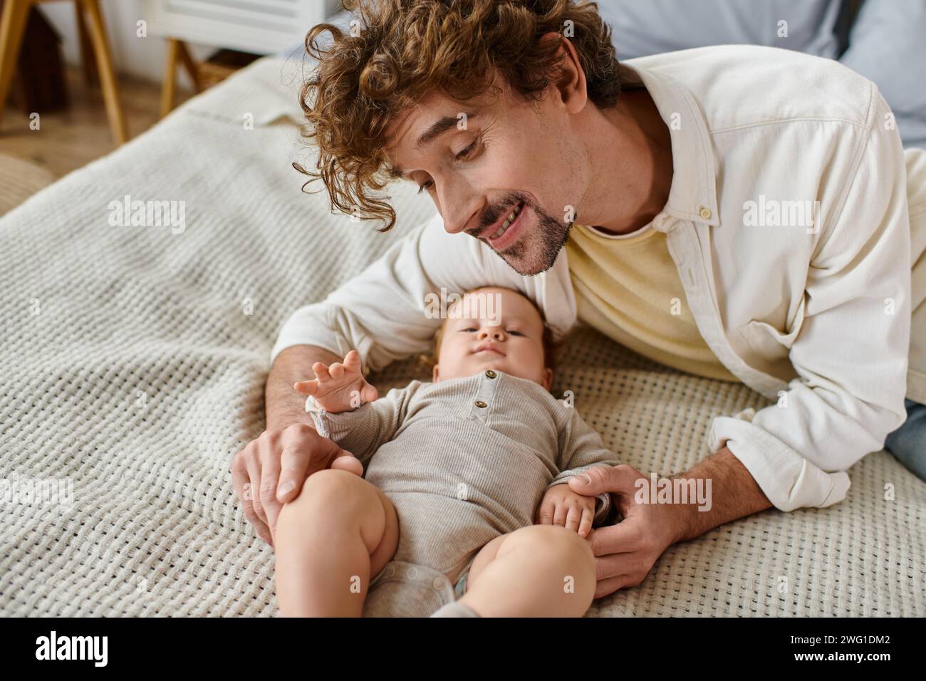 Fröhlicher Mann mit lockigen Haaren und Bart, der auf den süßen Baby-Sohn im Bett blickt, kostbare Momente Stockfoto