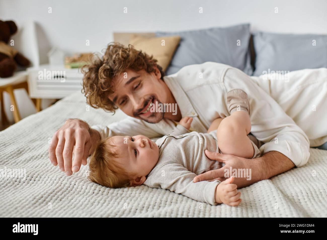 Fröhlicher Vater mit lockigen Haaren und Bart, der seinen kleinen Sohn im Bett ansieht, kostbare Momente Stockfoto