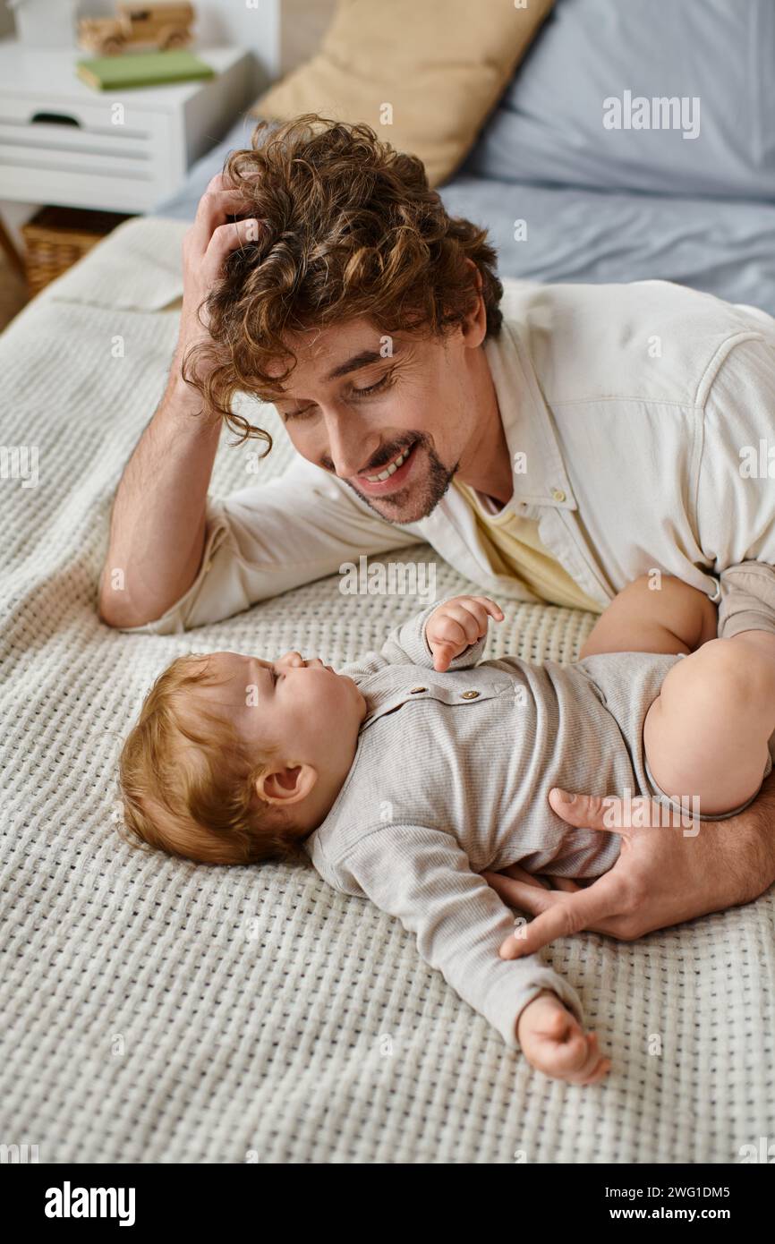 Fröhlicher Mann mit lockigen Haaren und Bart, der seinen kleinen Jungen im Bett ansieht, kostbare Momente Stockfoto