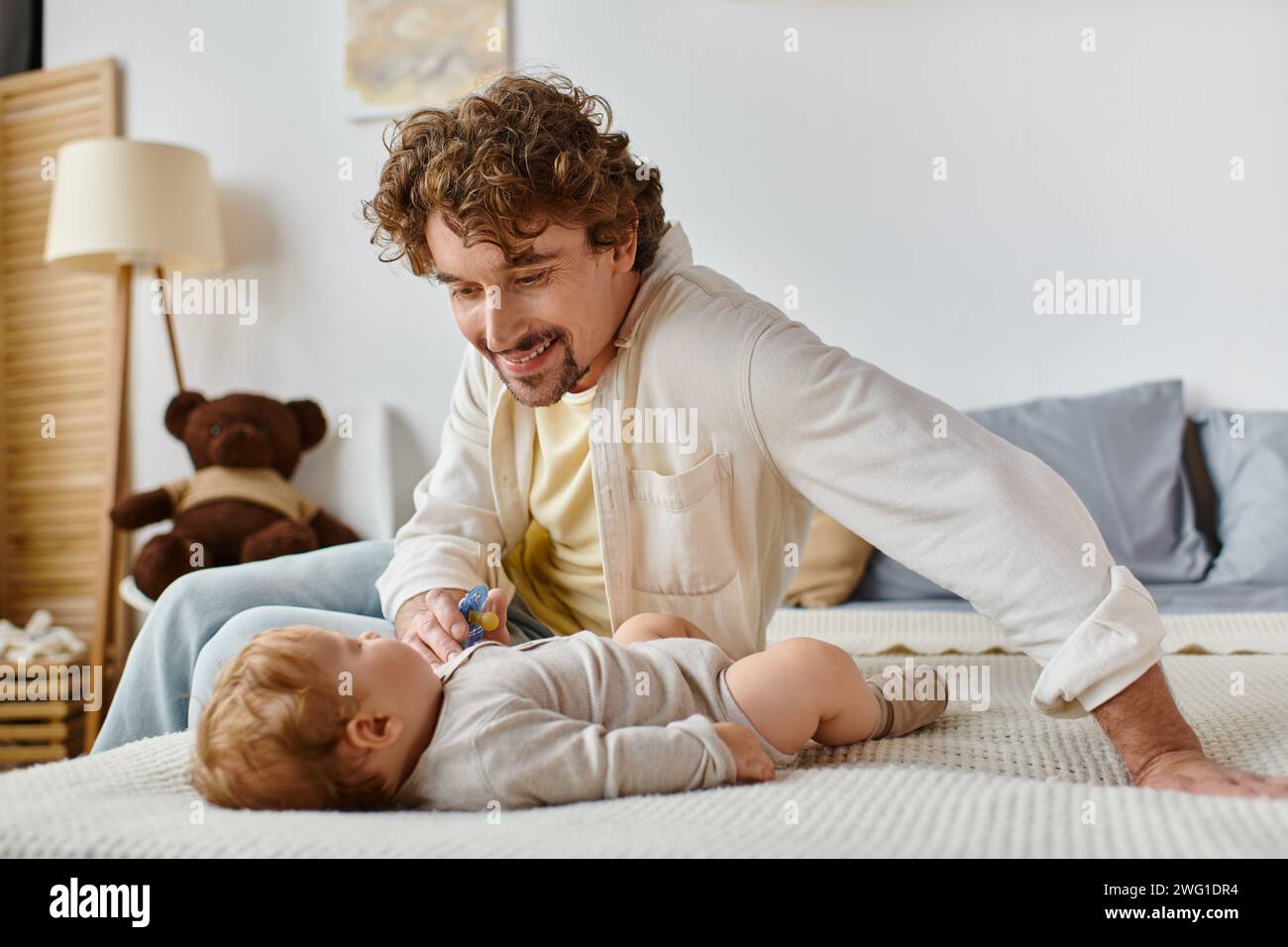 Fröhlicher alleinerziehender Vater, der seinem kleinen Sohn auf dem Bett Schnuller gibt, Vaterschaft und Liebe Stockfoto