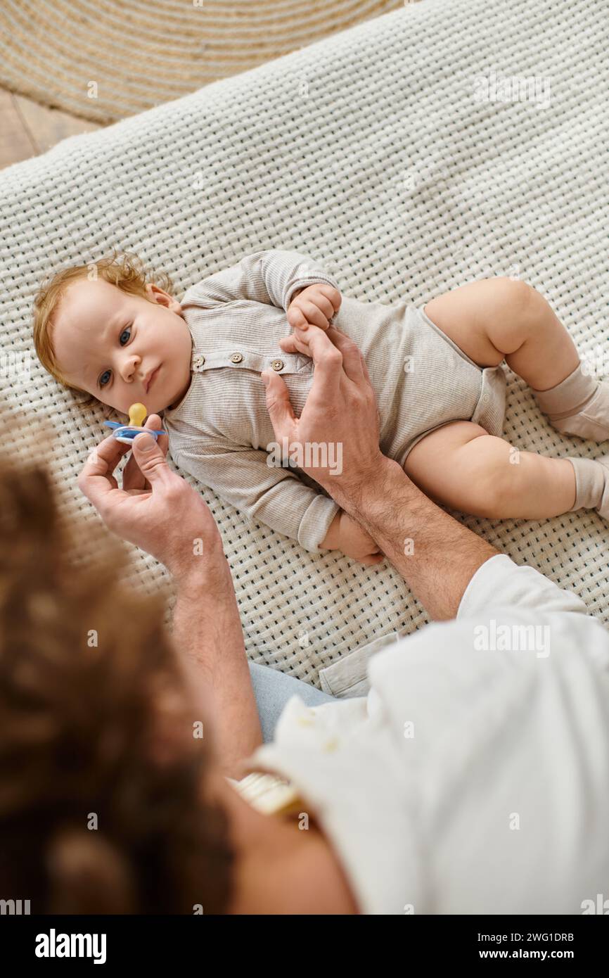 Blick von oben auf das Baby, das seinen verschwommenen Vater mit Schnuller in der Hand, Vaterschaft und Liebe betrachtet Stockfoto