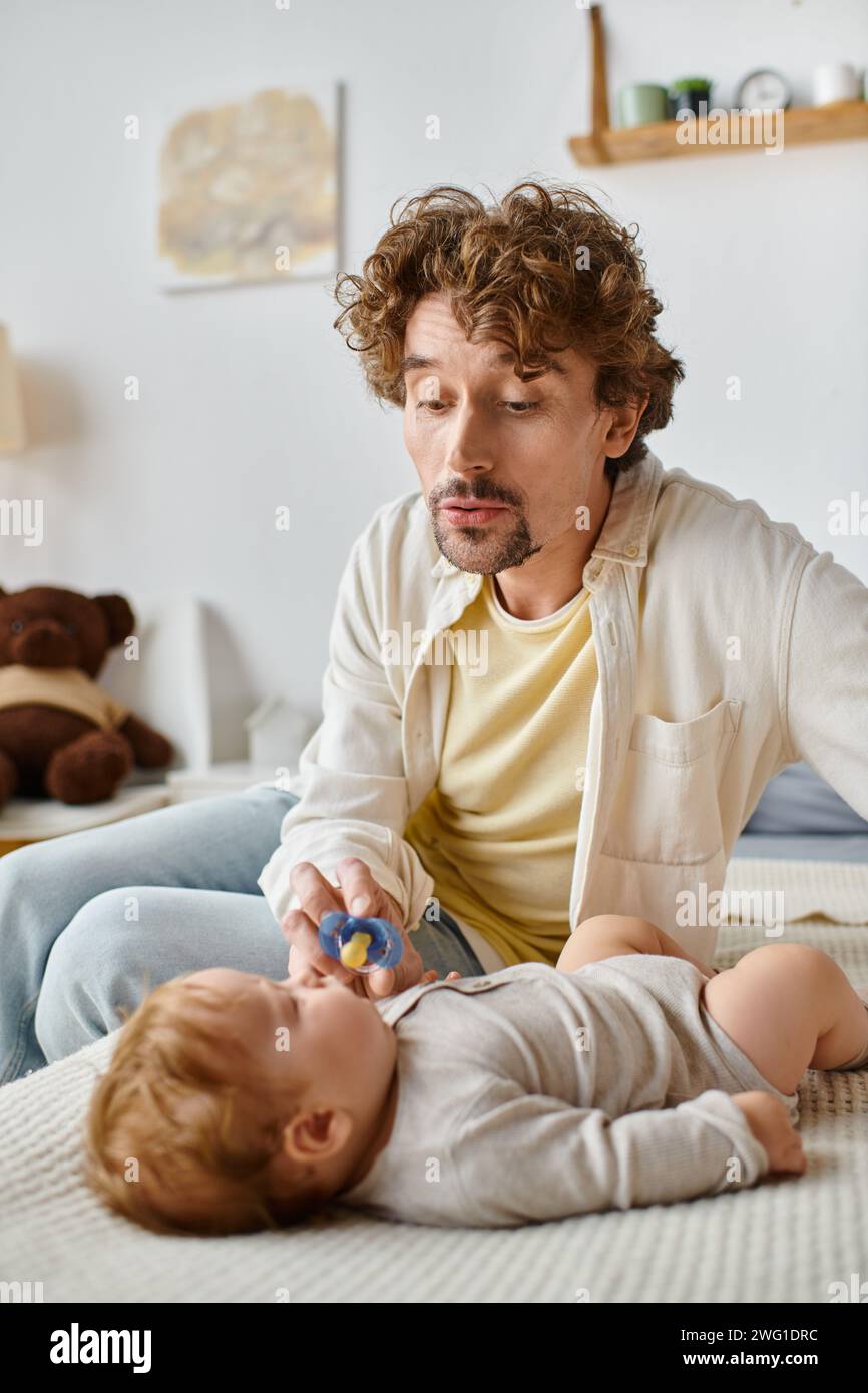 Alleinerziehender Vater macht komisches Gesicht, während er seinem kleinen Sohn auf dem Bett einen Schnuller gibt, Vaterschaft Stockfoto