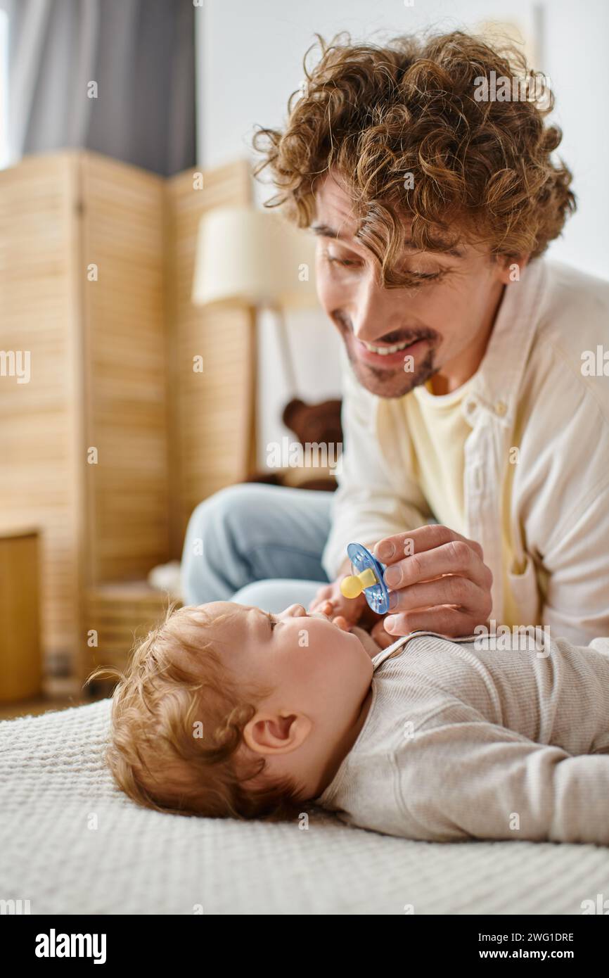 Glücklicher und lockiger Vater, der seinem kleinen Sohn auf einem gemütlichen Bett einen Schnuller schenkt, Vaterschaft und Liebe Stockfoto