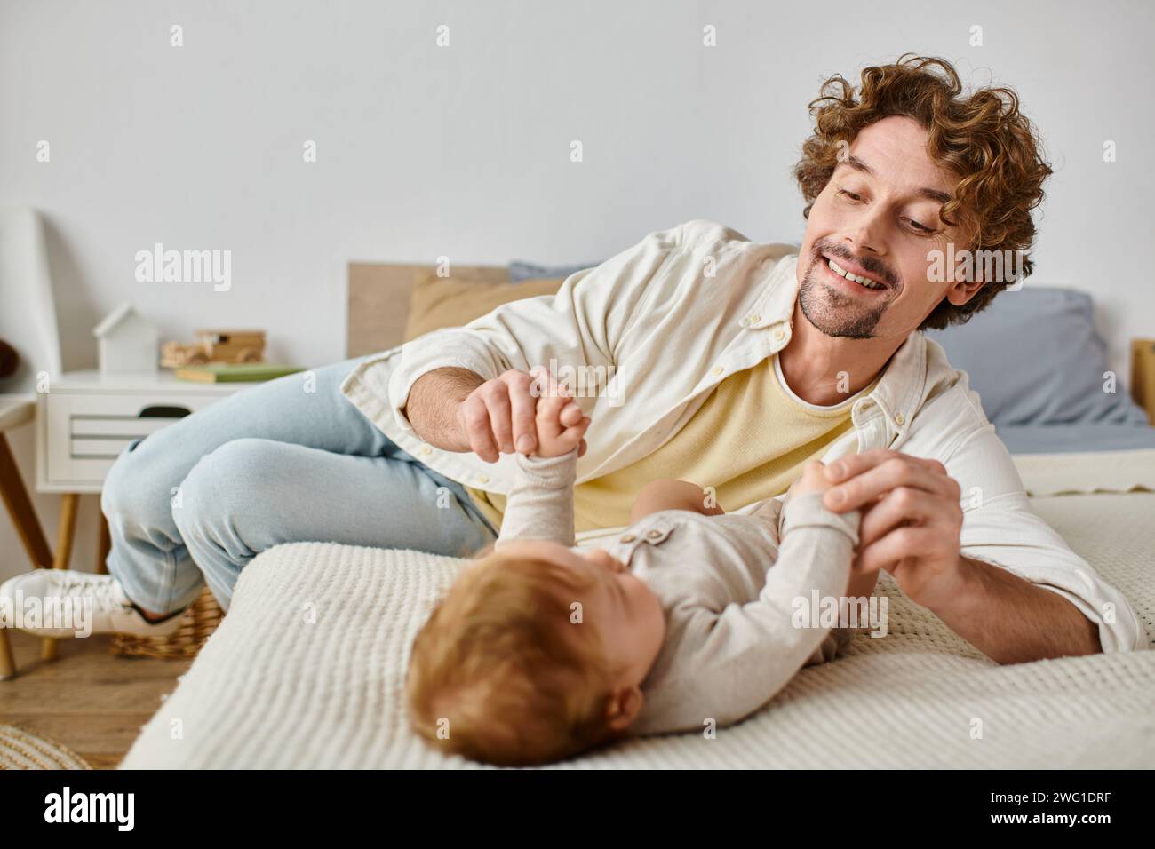 Glücklicher Vater mit lockigen Haaren, der die Hände seines kleinen Jungen hält, während er zusammen auf dem Bett lag, unschuldig Stockfoto