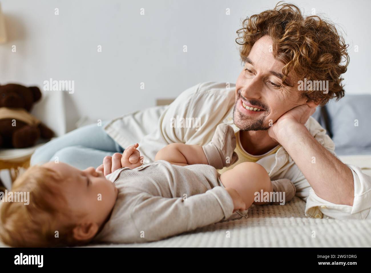 Fröhlicher Vater mit lockigen Haaren und Bart, der seinen kleinen Sohn ansieht, während er zusammen auf dem Bett liegt, Liebe Stockfoto