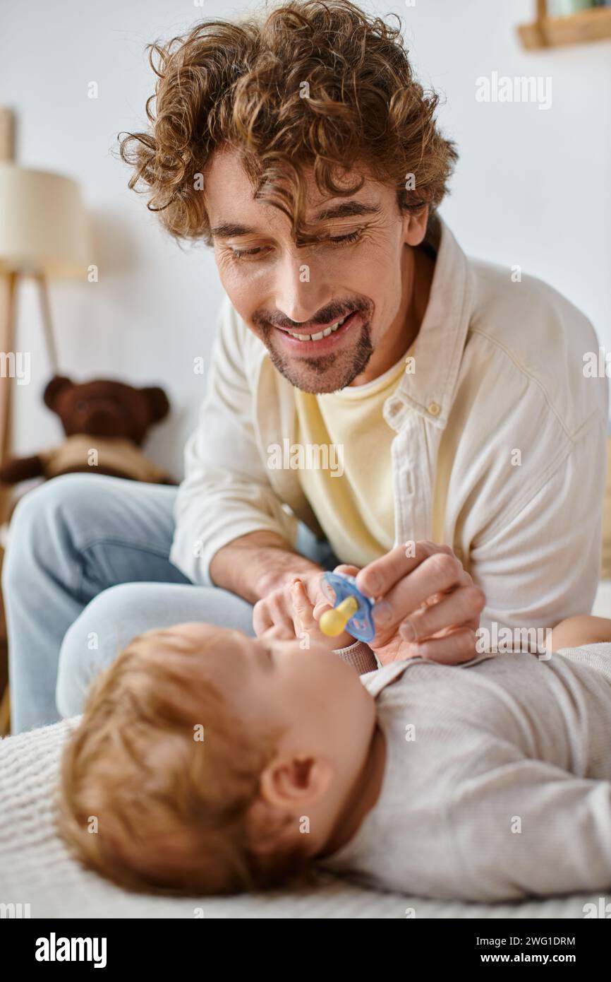 Fröhlicher und lockiger Vater, der seinem kleinen Sohn auf dem Bett Schnuller gibt, Vaterschaft und Liebe Stockfoto