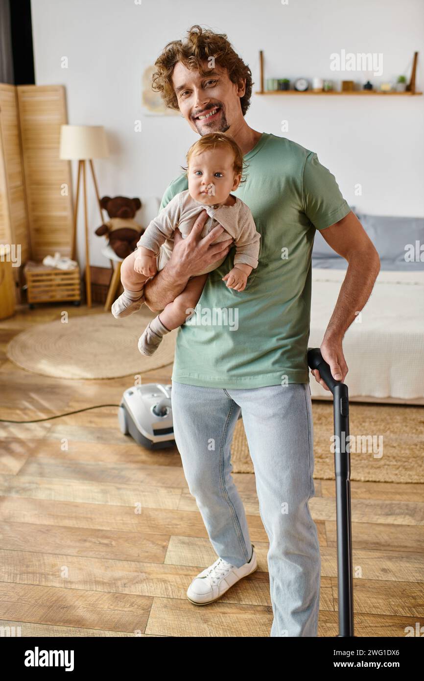 Glücklicher Mann, der Hausarbeit und Kinderbetreuung betreibt, Vater saugt Hartholzboden mit Sohn in Armen Stockfoto