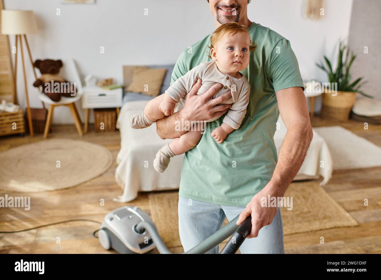 Ein zugeschnittener Mann, der Hausarbeit und Kinderbetreuung betreibt, ein glücklicher Vater, der mit einem Sohn in den Armen ein Haus saugt Stockfoto