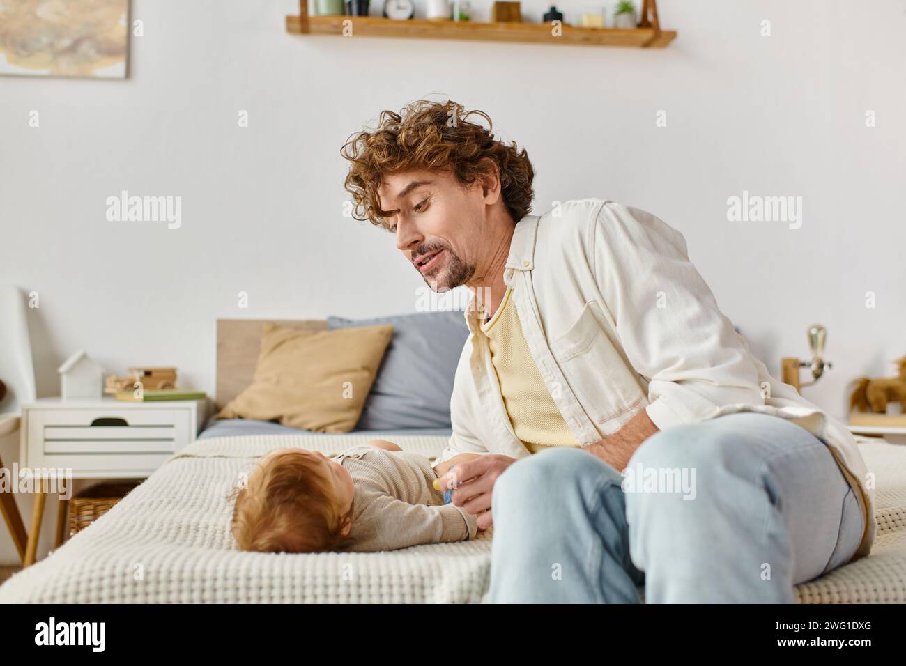 Alleinerziehender Vater mit lockigen Haaren, der einen Schnuller in der Nähe des kleinen Jungen auf dem Bett hält, Vaterschaft und Liebe Stockfoto