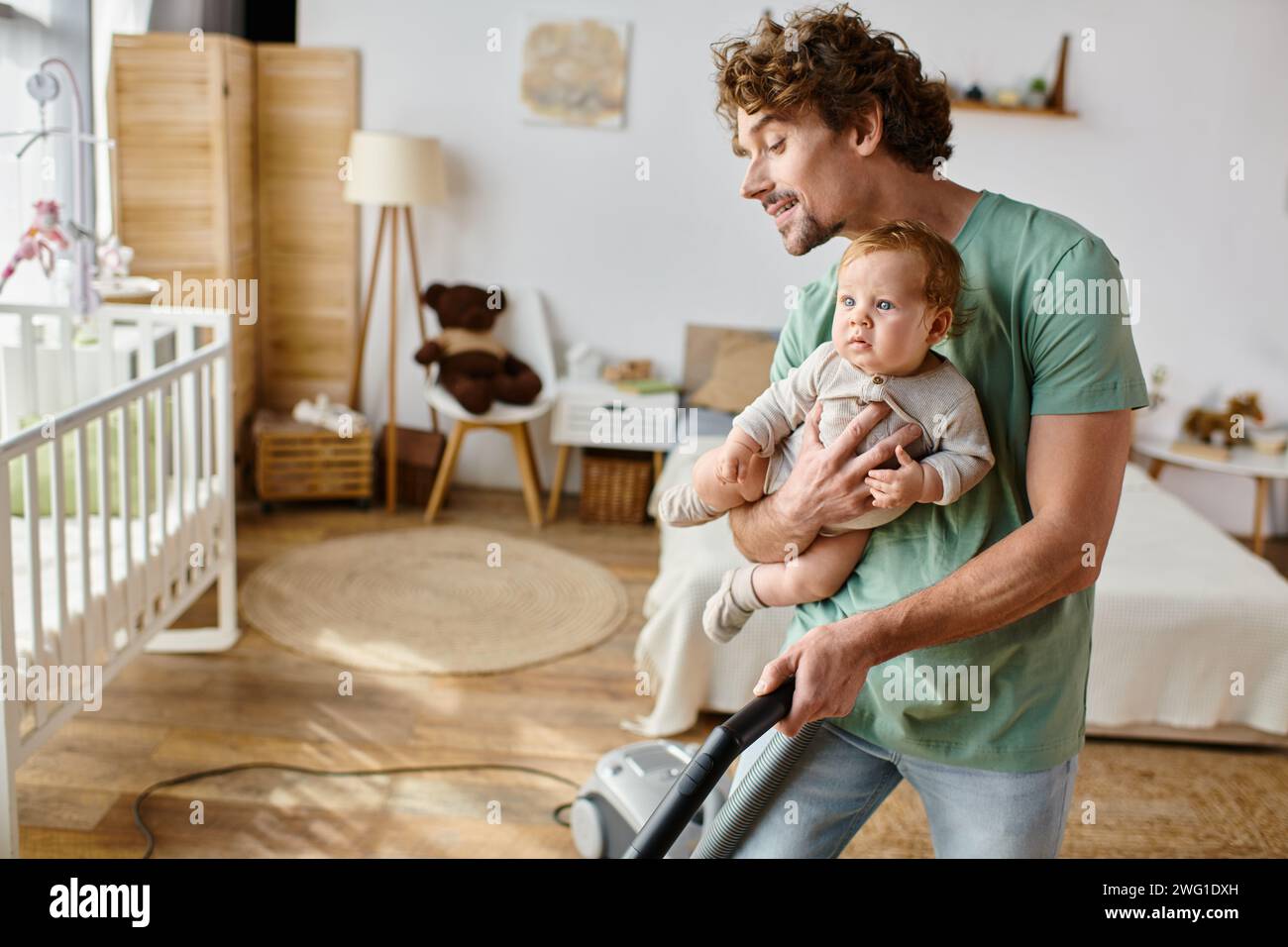 Mann Multitasking Hausarbeit und Kinderbetreuung, Vater saugt Hartholzboden mit dem kleinen Sohn in den Armen Stockfoto