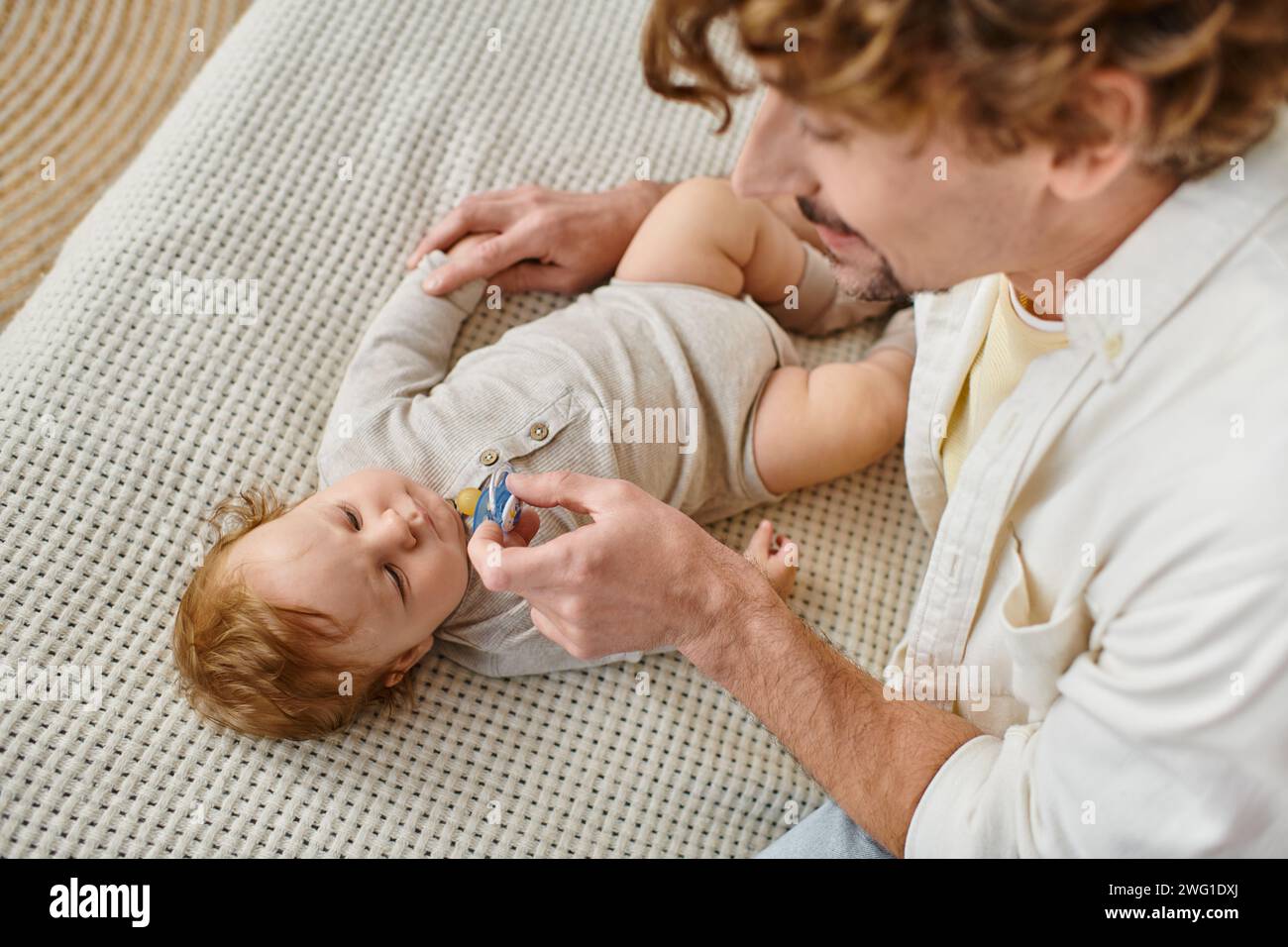 Blick von oben auf den lockigen und bärtigen Vater, der einen Schnuller in der Nähe des kleinen Jungen auf dem Bett hält, Vaterschaft Stockfoto
