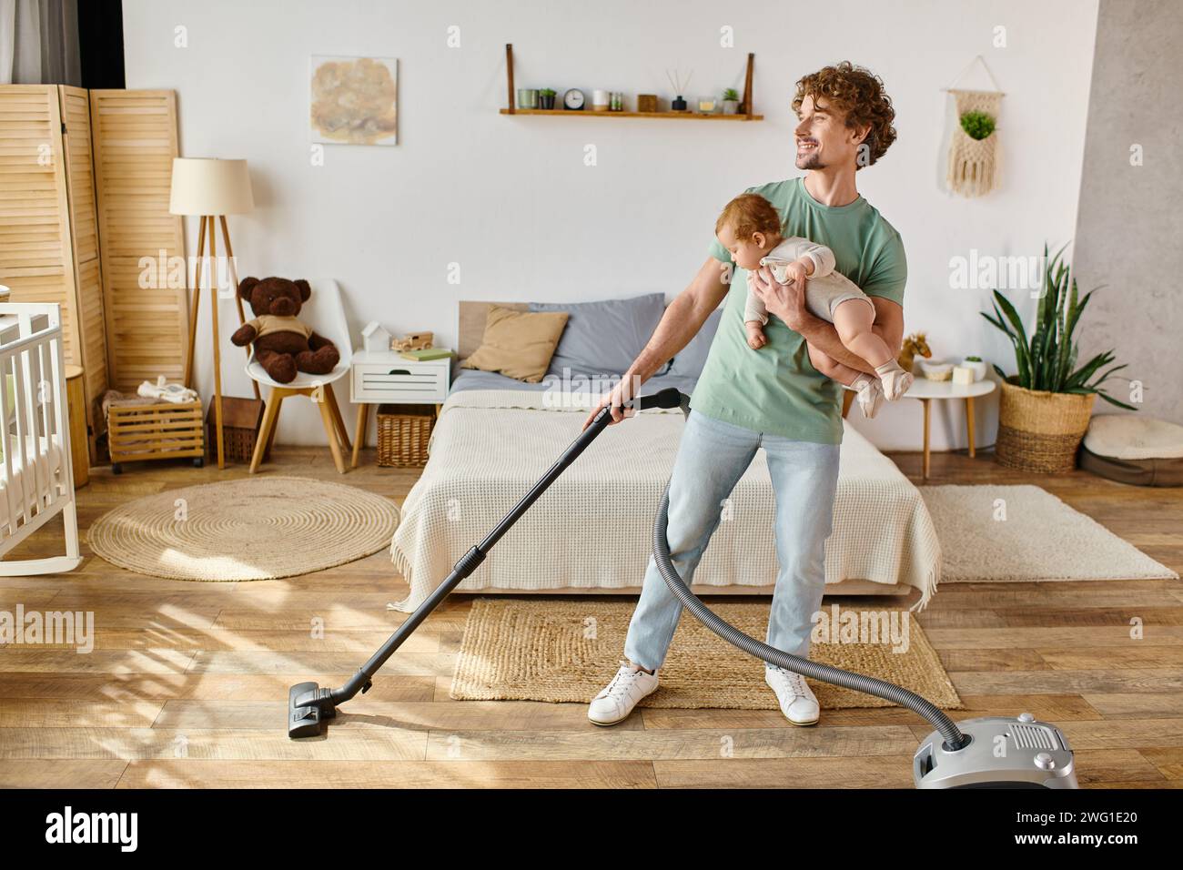 Glücklicher Mann, der Hausarbeit und Kinderbetreuung betreibt, Vater saugt das Schlafzimmer mit einem Jungen in Armen Stockfoto
