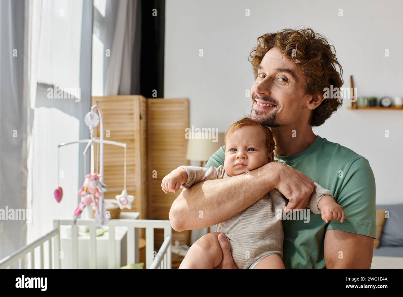 Lockiger und glücklicher Vater, der seinen kleinen Sohn in den Armen hält, in einem gemütlichen Schlafzimmer zu Hause, Vaterschaft Stockfoto