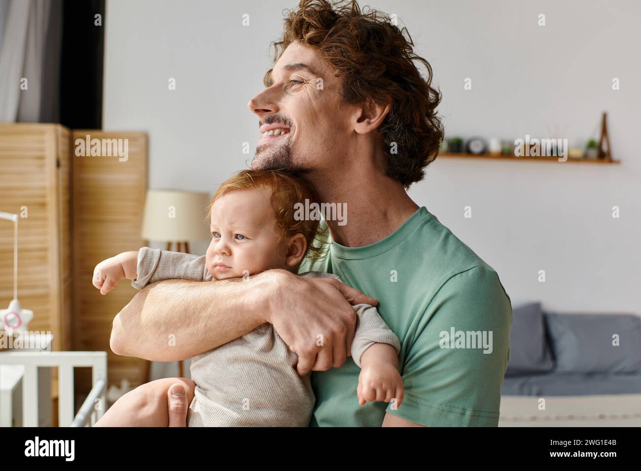 Lockiger und fröhlicher Vater, der seinen kleinen Sohn in den Armen hält, in einem gemütlichen Schlafzimmer zu Hause, Vaterschaft Stockfoto