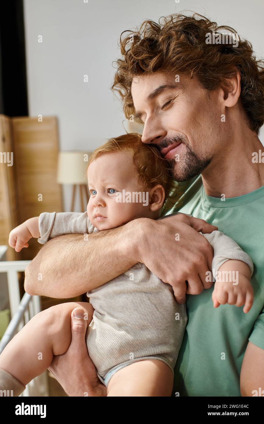 Lockiger und fröhlicher Vater, der seinen kleinen Sohn in den Armen hält, in einem gemütlichen Schlafzimmer zu Hause, Vaterschaft Stockfoto