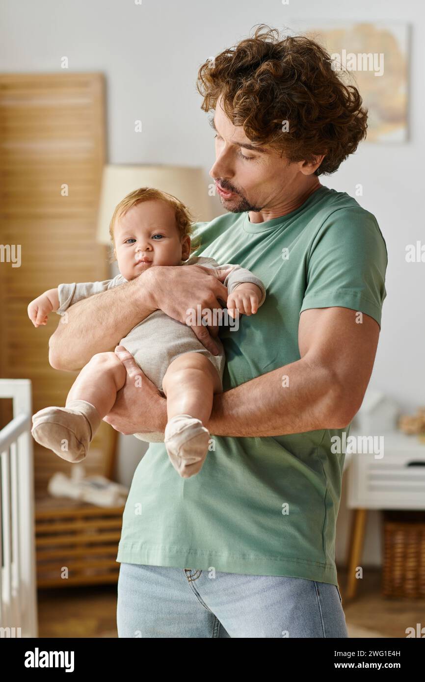 Lockiger und bärtiger Mann, der seinen kleinen Sohn in den Armen hält, in einem gemütlichen Schlafzimmer zu Hause, Vaterschaft Stockfoto