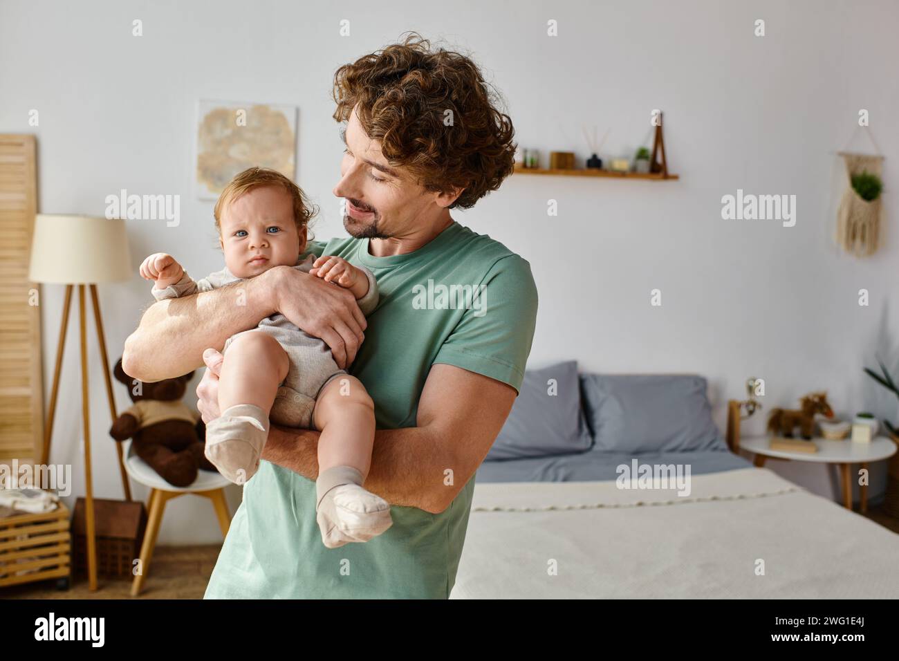 Lockige und glückliche Vater hält seinen kleinen Jungen in einem gemütlichen Schlafzimmer zu Hause, Vaterschaft Stockfoto