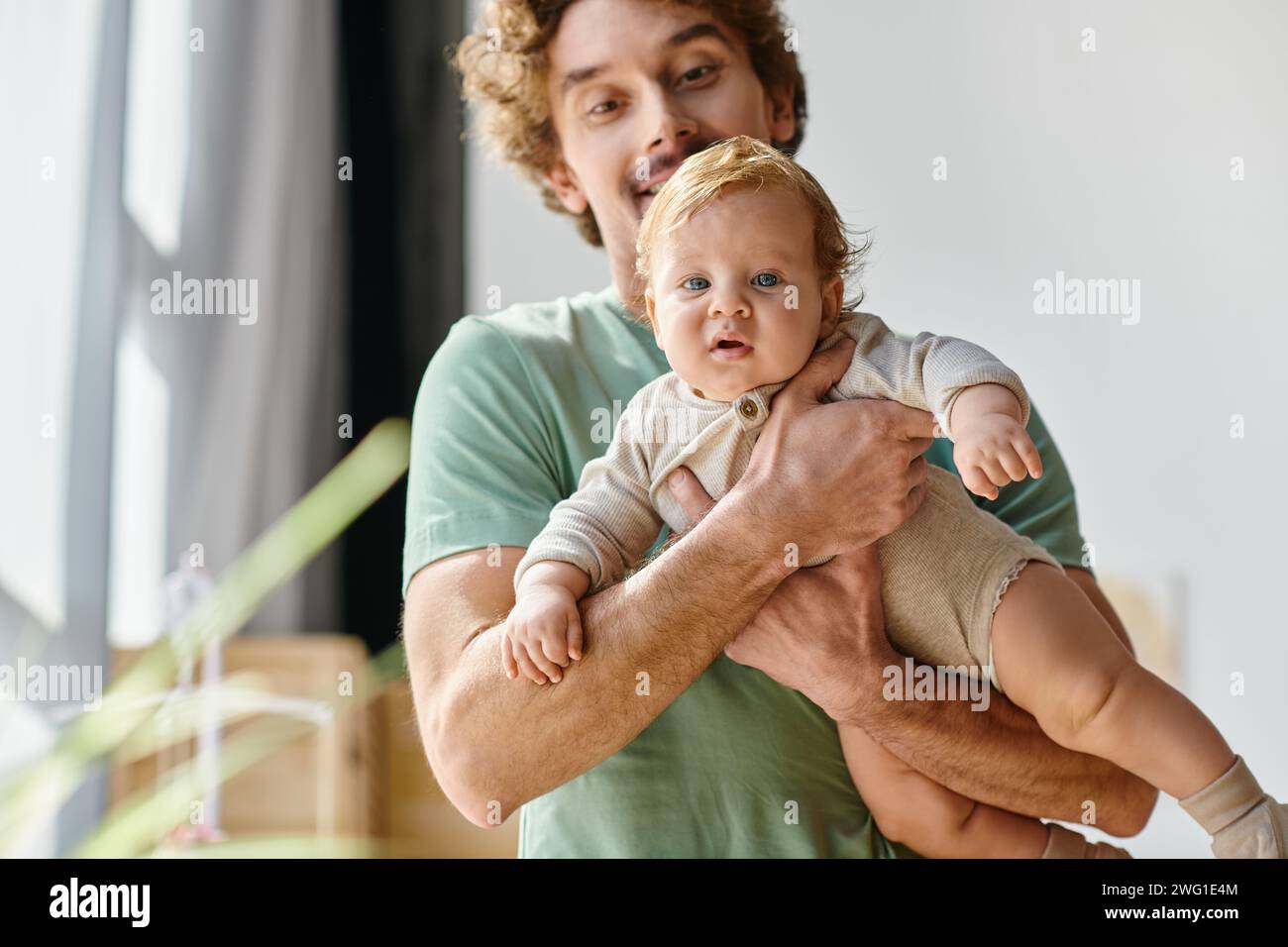 Lockiger und bärtiger Mann, der seinen kleinen Sohn mit blauen Augen zu Hause in den Armen hält, Vaterschaft Stockfoto