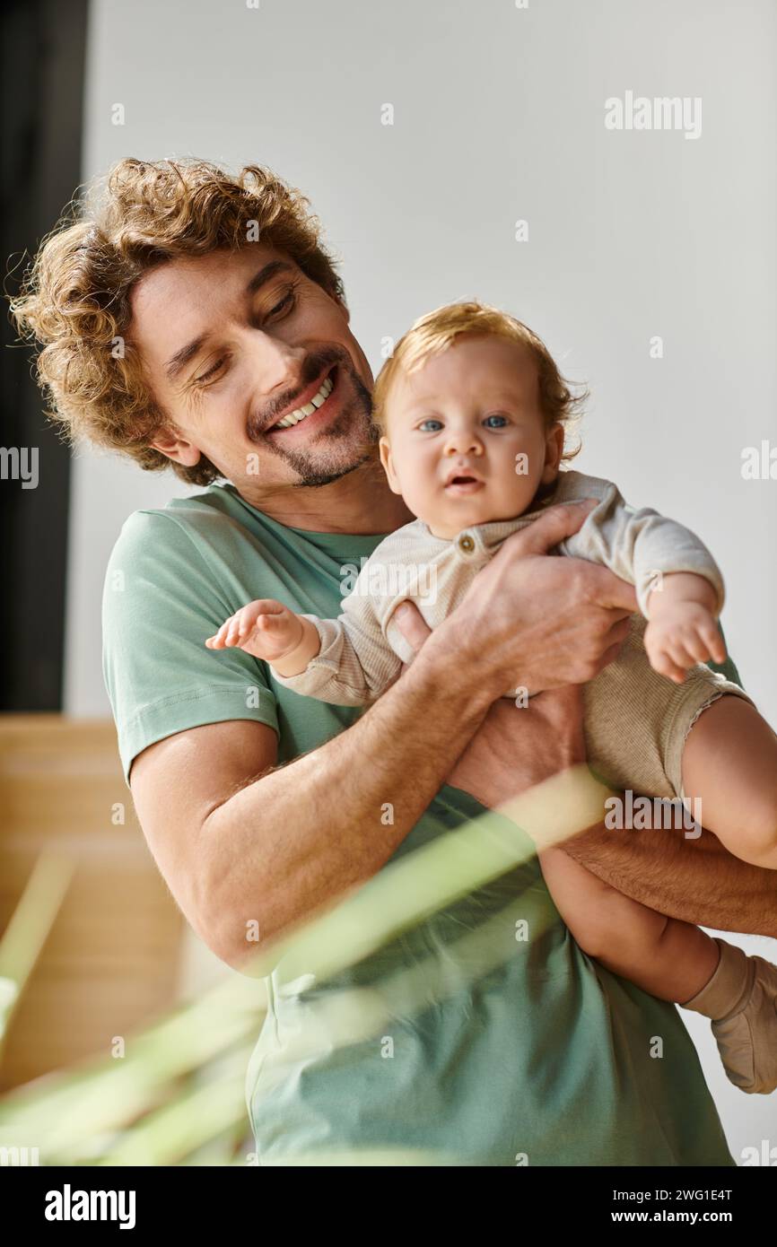 Lockiger und zufriedener Vater, der seinen kleinen Sohn in den Armen hält, in einem gemütlichen Schlafzimmer zu Hause, Vaterschaft Stockfoto