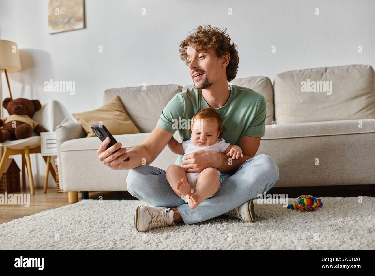 Glücklicher alleinerziehender Vater mit Smartphone, während er mit dem Kleinkind auf dem Teppich in der Nähe von Rassel sitzt Stockfoto