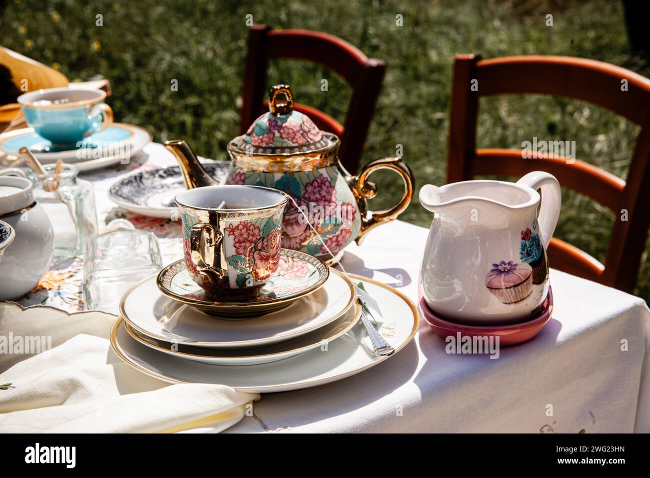 „Erleben Sie die Eleganz einer klassischen Teezeit mit dieser exquisiten Teekanne mit Blumenmuster und einer passenden Tasse, die auf einem sonnigen Tisch mit pastoralem Charme steht.“ Stockfoto